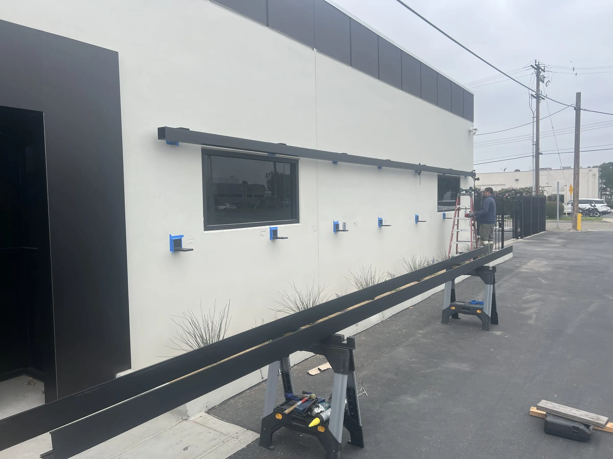 Installer on a ladder fixing black facade components to a white wall, more components on sawhorses in front