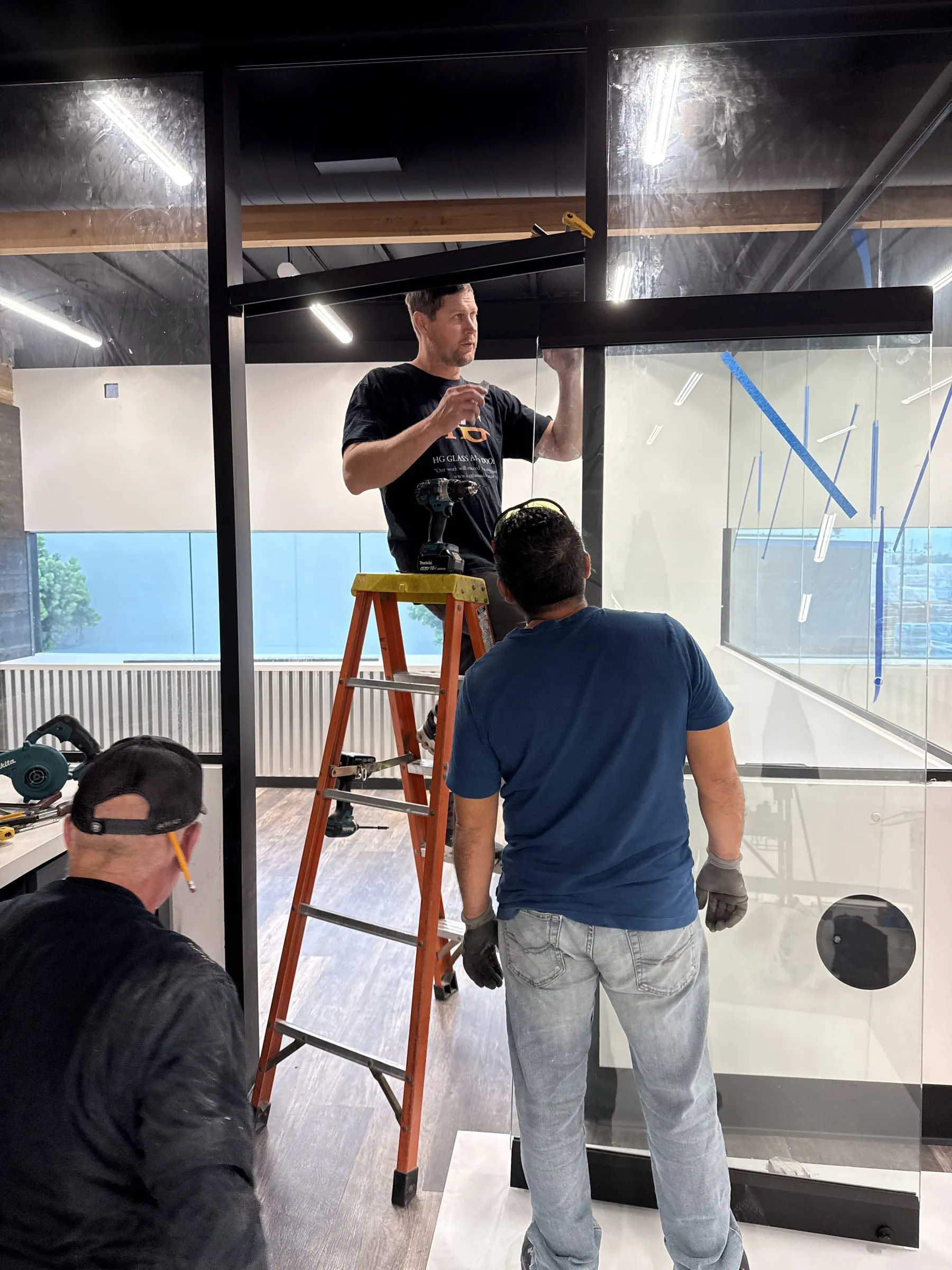 Three installers fitting a glass door in black-framed office partitions, one on a ladder in an HG Glass and Doors shirt