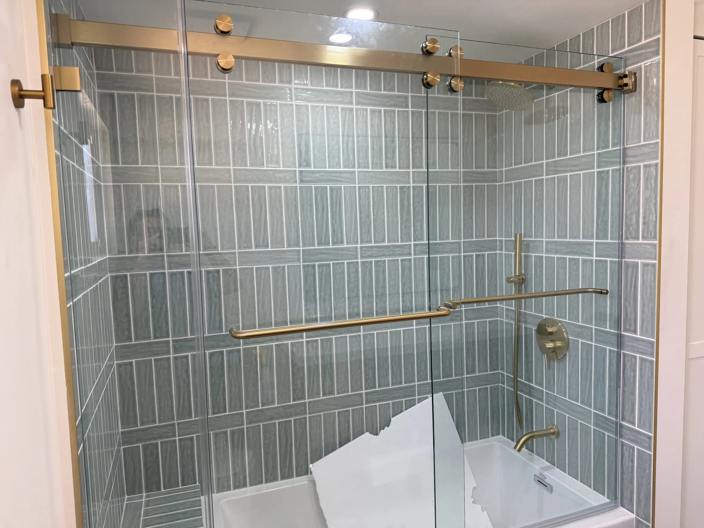 Brass track and rollers of frameless sliding glass tub doors against green-gray tile