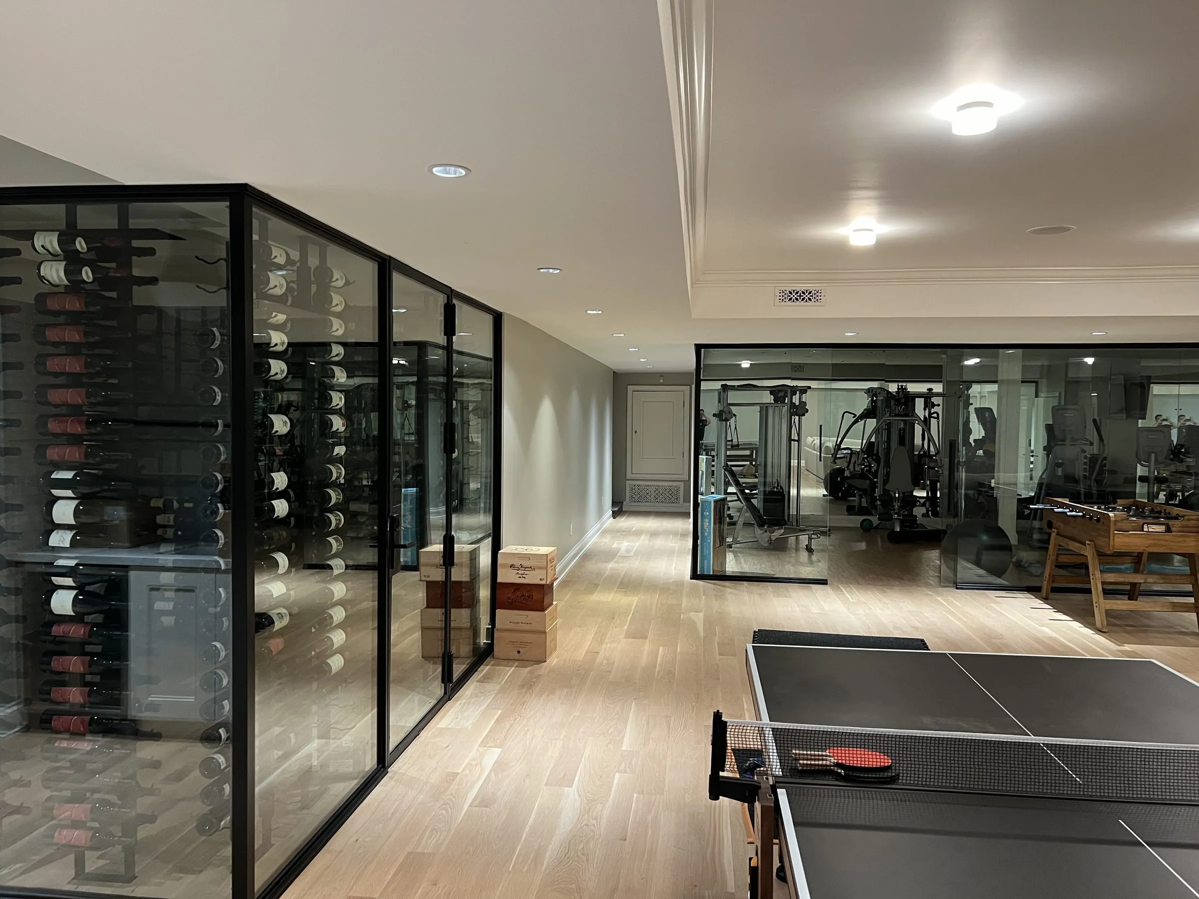 Black-framed glass wine cellar in the foreground and the glass-walled home gym behind it, across a games room